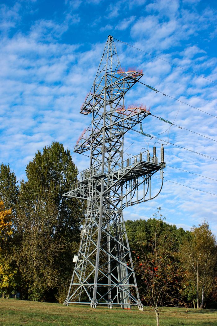 Home A towering electricity pylon set against a clear blue sky and lush greenery, symbolizing power and nature.